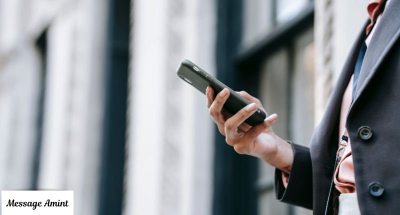 A person in a business suit holding a smartphone outdoors, typing a text message with a blurred city background. "Message Amint" text logo is visible in the bottom-left corner.